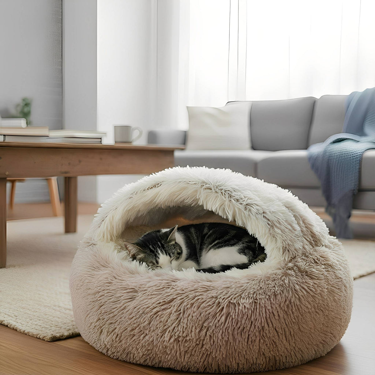 Zwart-witte kat ligt opgerold in beige kattenmand naast een grijze bank